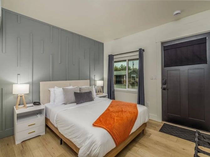 Full-bed studio at The Flats: white linens, grey accent wall, orange throw blanket