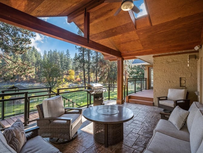 Covered outdoor living area at The River House with fire pit table, grill, and Spokane River views through the trees